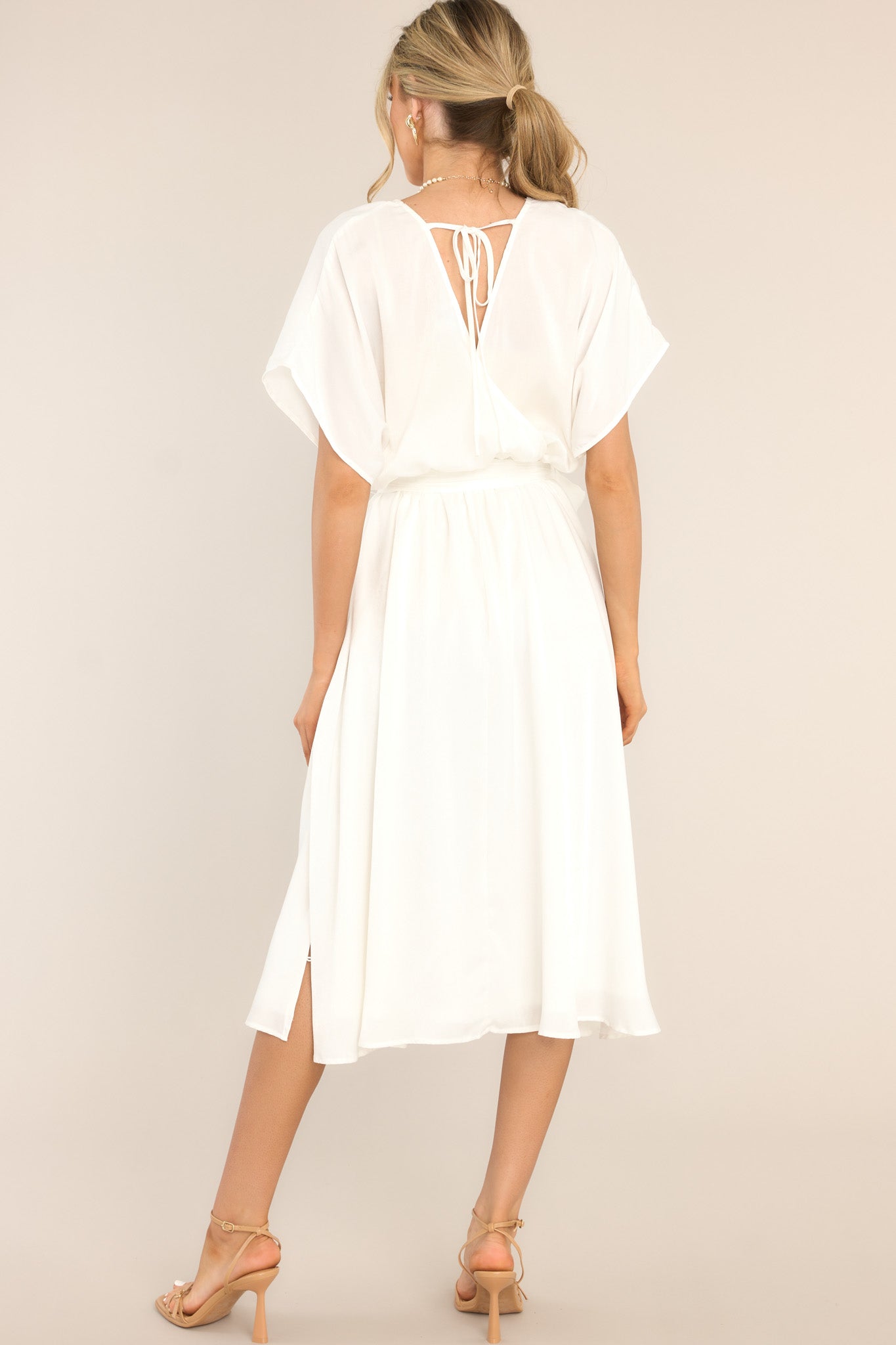 Back view of this midi dress that features a V-neckline, an open back with a self-tie closure, flowy cap sleeves, an elastic waistband with a self-tie fabric belt, and button detailing down the side of the skirt.
