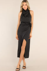 This all black dress features a high cowl neckline, a zipper down the back, a wrap-like design with a self-tie feature at the waist, and a front slit. 