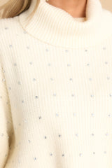 Close up of rhinestones on an ivory knit sweater featuring a turtle neckline and ribbed texture. 