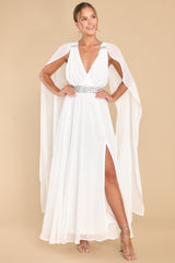 This white dress features a v-neckline, adjustable crisscross straps down the back, rhinestone detailing, fabric trailing from the shoulder to the bottom of the dress, and a slit on the left side of the skirt.