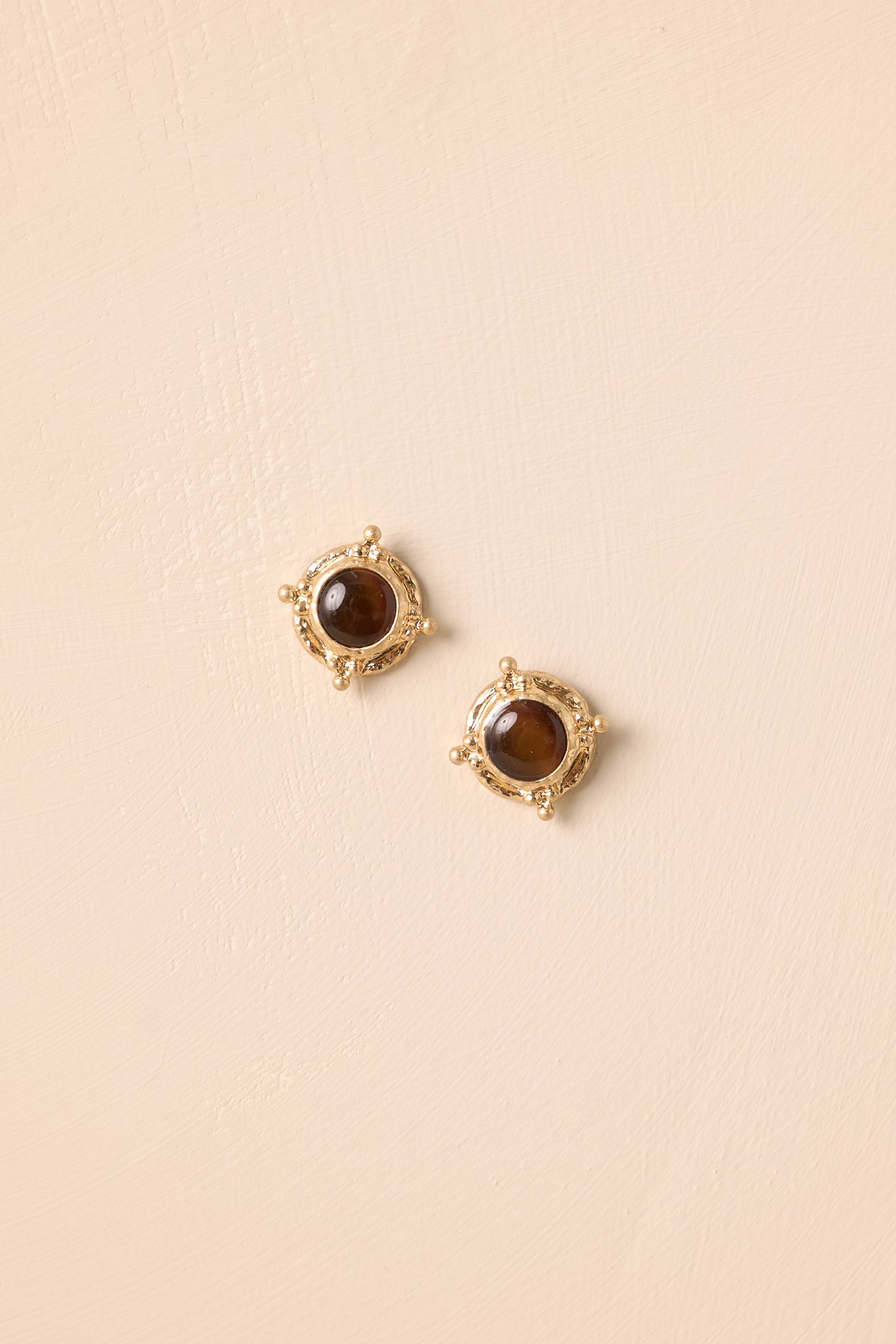 Close-up view of round stud earrings with glossy brown cabochon centers framed in textured gold with small bead accents on beige background.