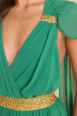 Close up of this dress that features a deep v-neckline, gold rhinestone detailing around the collar bone area and waistline, fabric trailing from the collarbone towards the back of the shoulder to the bottom of the dress.