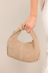 This taupe bag features a short handle with a knotted detail, a removable gold chain strap that is non-adjustable, a zipper closure, a zipper pocket on the inside, and a suede finish.