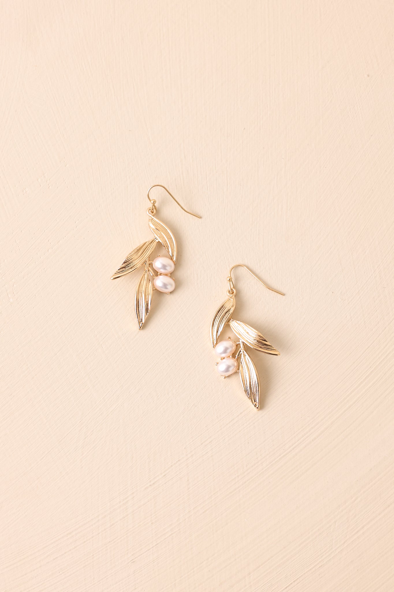 Flat lay of the earrings on a beige surface emphasizing the delicate leaf design and two ivory faux pearls.