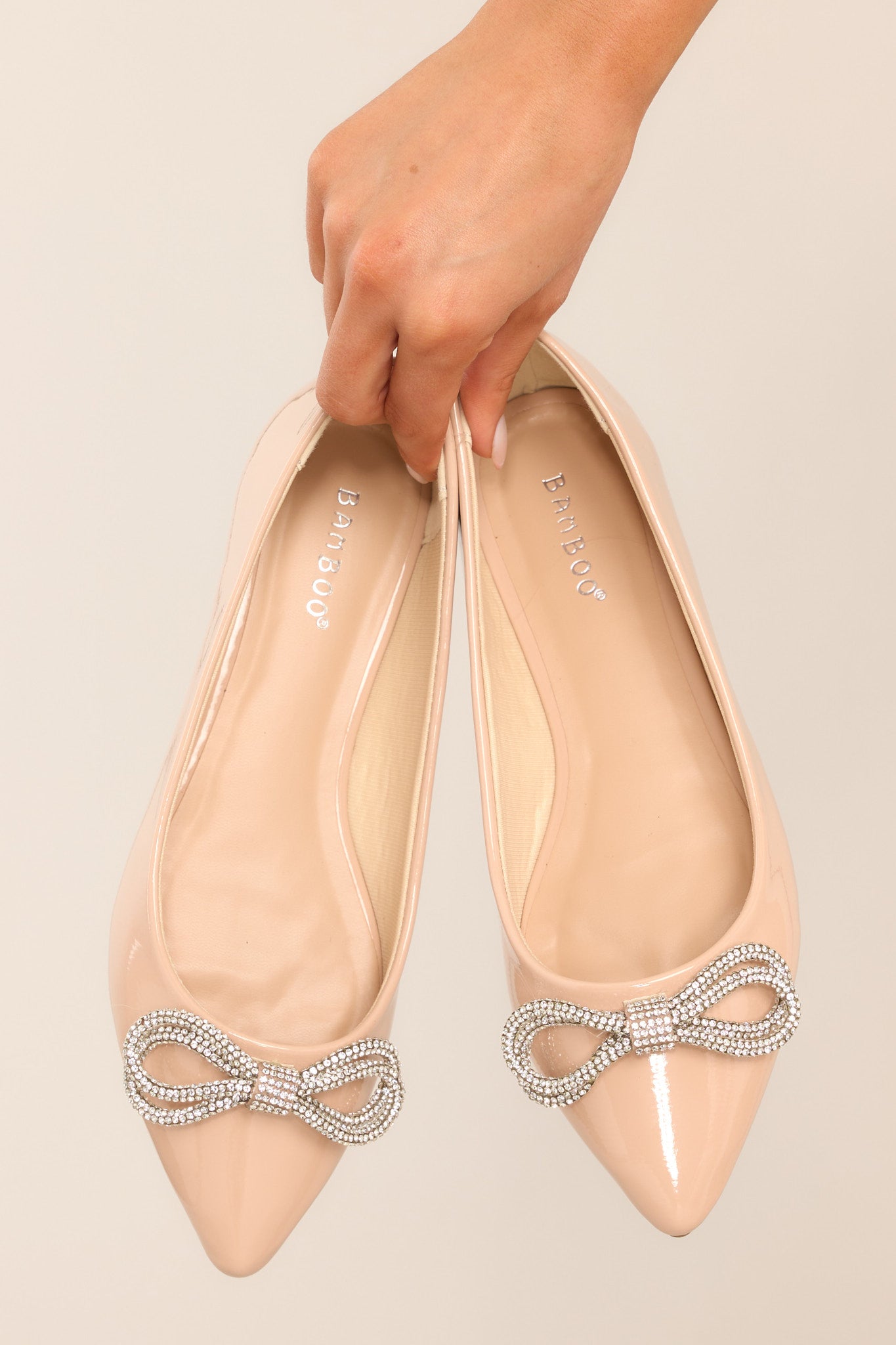 These nude colored flats feature a pointed toe, a rhinestone embellished bow, and a shiny finish. 