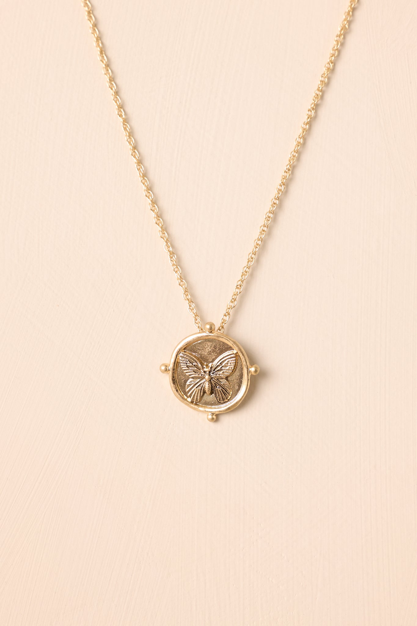 Gold medallion necklace displayed against a beige background, featuring the round pendant with detailed embossing and fine cable chain.