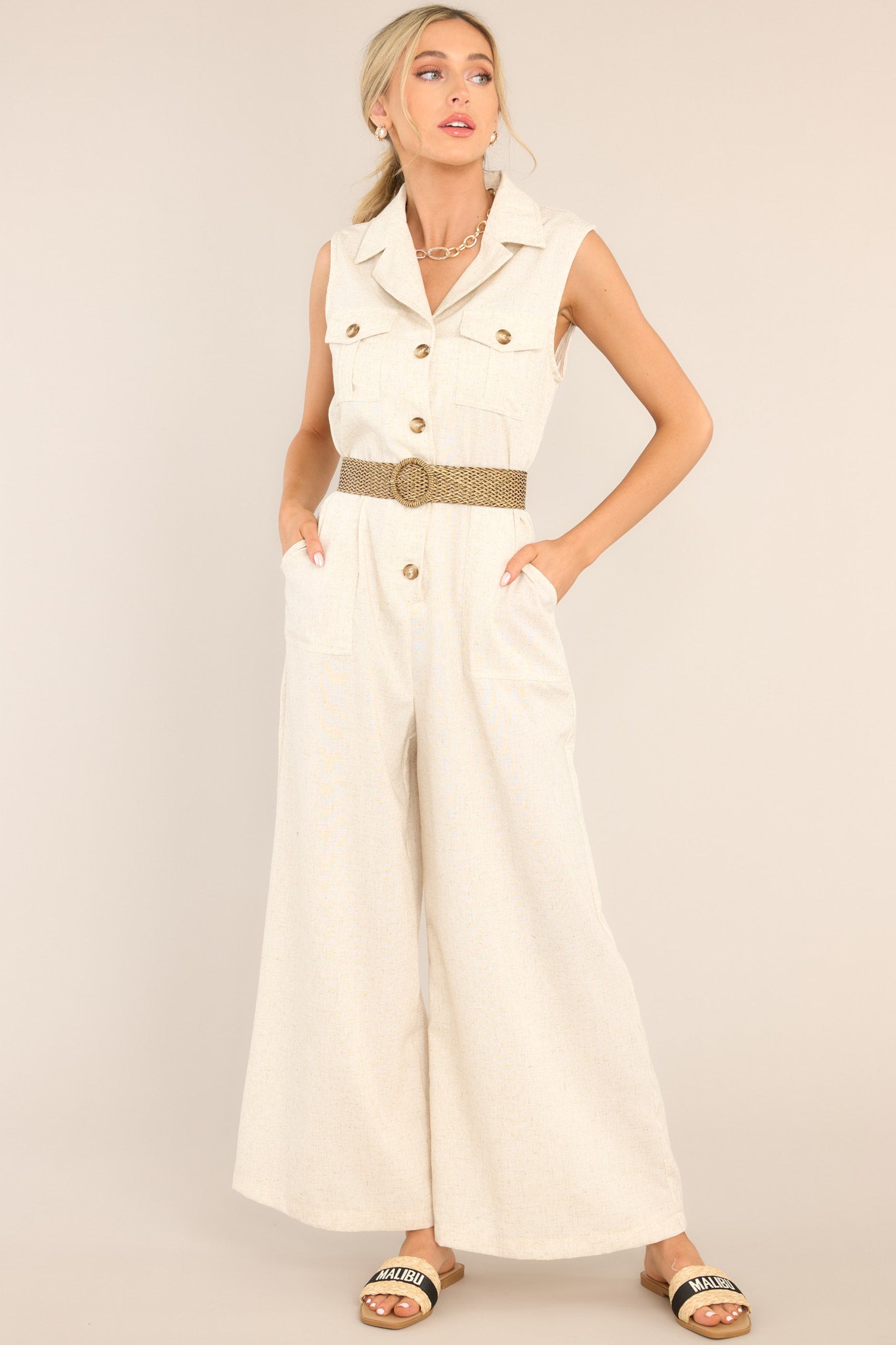 This oatmeal colored jumpsuit features a notched lapel collared neckline, chest pockets, a functional button front, belt loops, an adjustable belt, an elastic insert at the back of the waist, hip pockets, and a wide leg.