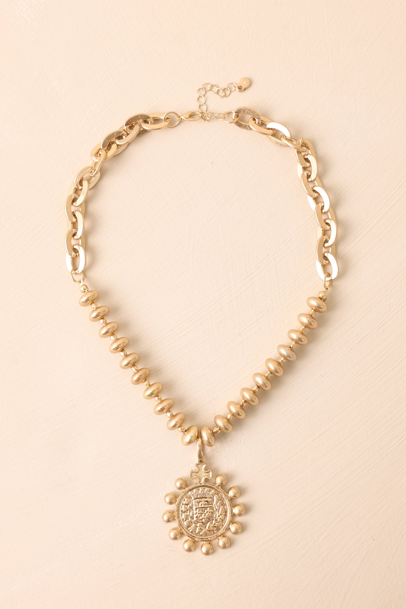 The layered chunky chain necklace shown against a beige backdrop featuring an embossed medallion and lobster claw clasp.