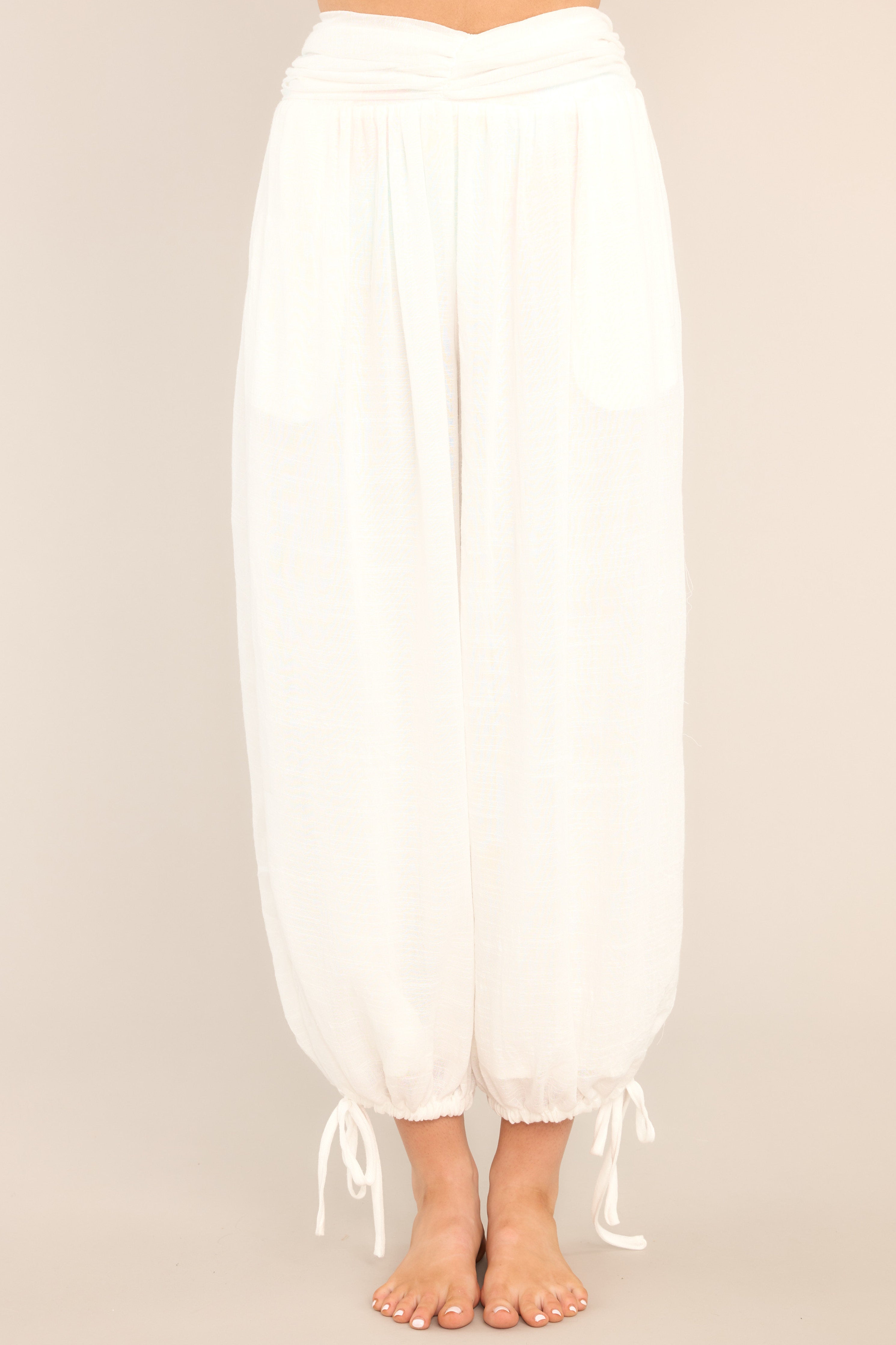 Front view of these pants that feature a high waisted design with cinching in the middle, a smocked insert in the back, functional hip pockets, a linen-like material, and self-tie drawstring cuffed ankles. 