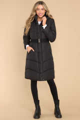 Puffed, knee length black coat featuring a high neckline and hood, a functional zipper down the front, an elastic belt, and pockets with zipper closures.