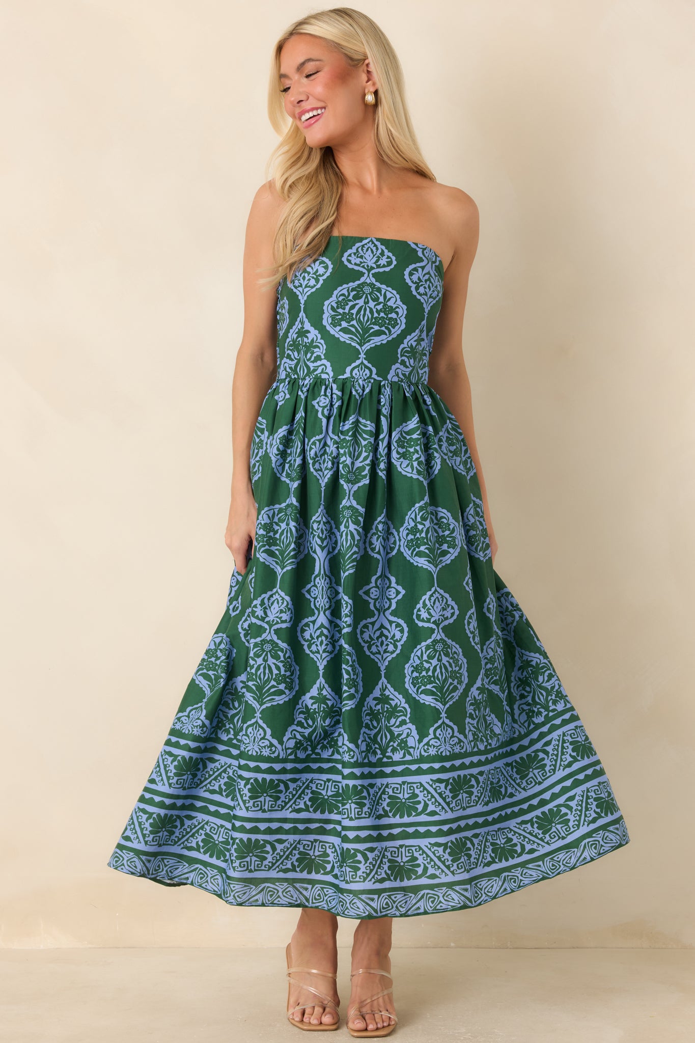 A woman modeling a strapless midi dress with a green and blue paisley pattern against a solid beige background.