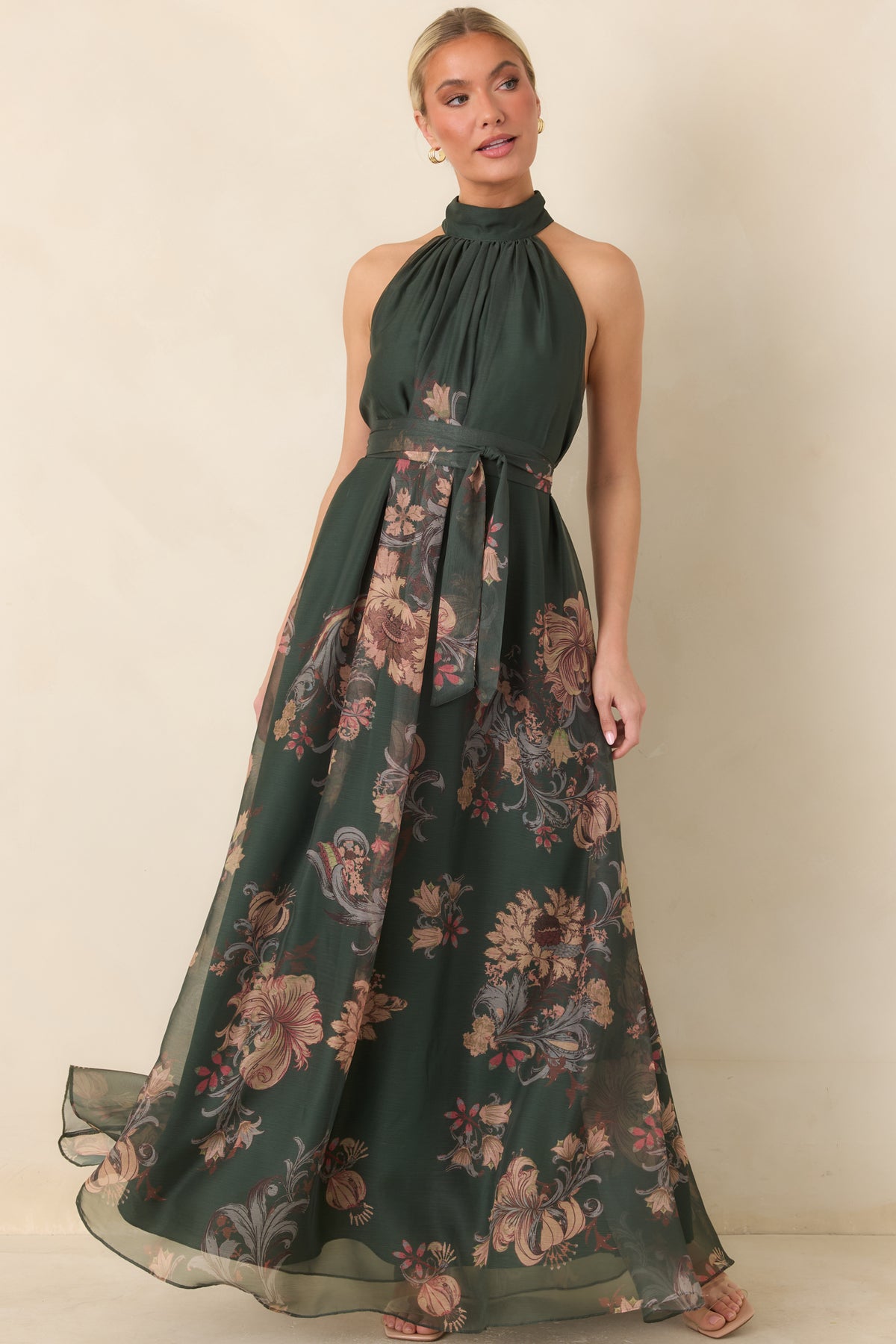 The forest green dress features a softly gathered halter bodice, defined waist sash, and layered maxi skirt with an airy finish.
