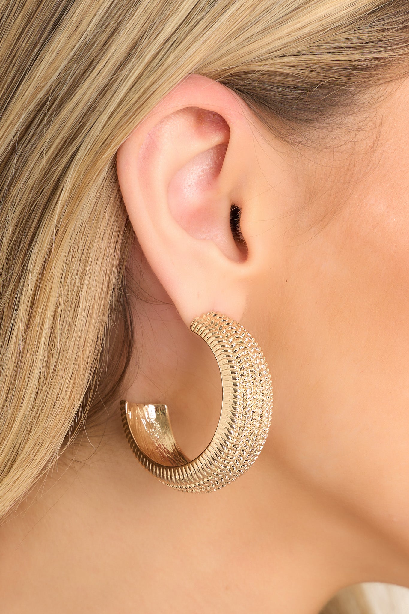 In-ear view of earrings that feature a gold textured finish, a wide hoop design, and secure post-back fastenings.