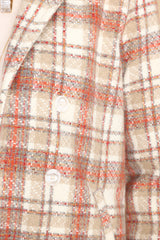 Close up view of this coat that features a collared neckline, double breasted buttons on the front, and a plaid pattern in shades of orange and beige.