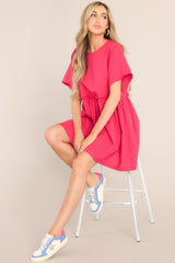 This pink dress features a round neckline, a keyhole cutout at the back of the neck with a button closure, short sleeves, ruffle detailing along the waistline, and functional pockets at the hips.