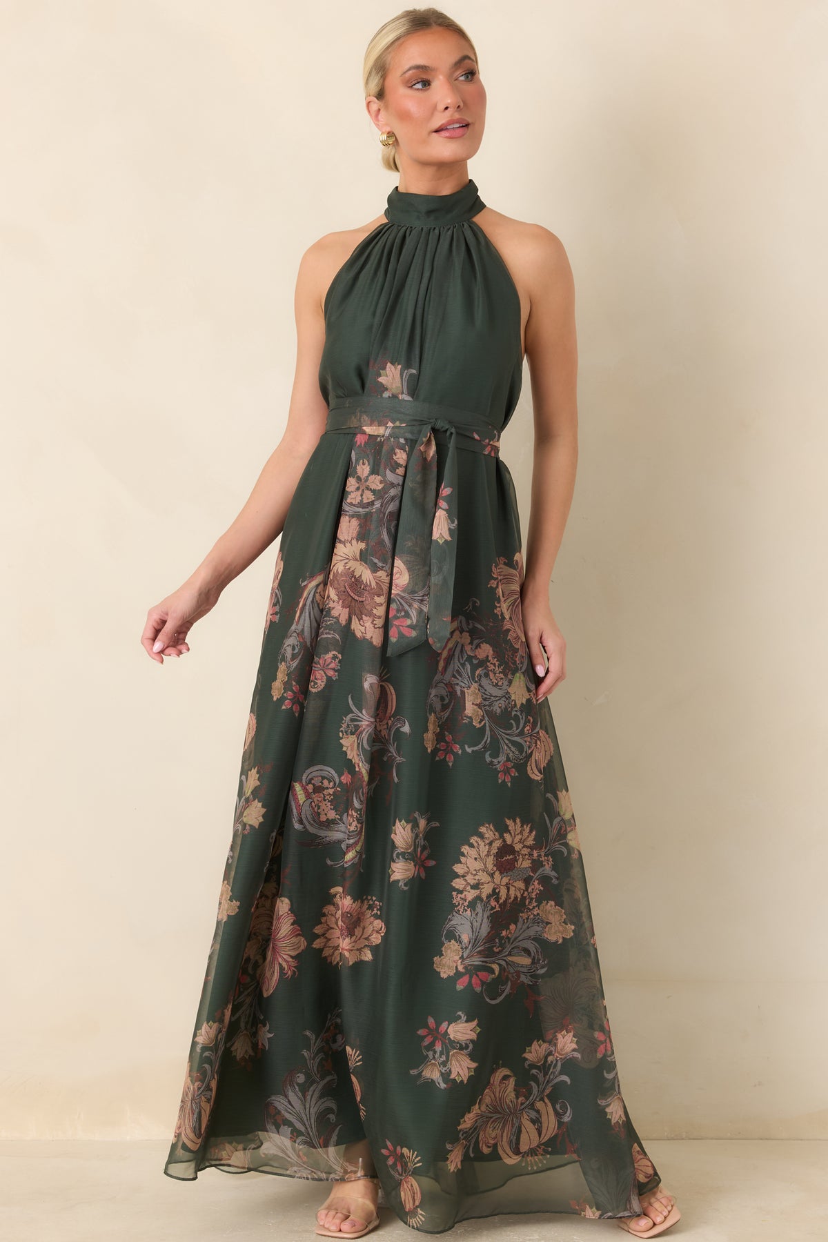 The forest green floral maxi dress features a coordinating waist sash and a softly draped halter bodice above its sweeping sheer skirt.