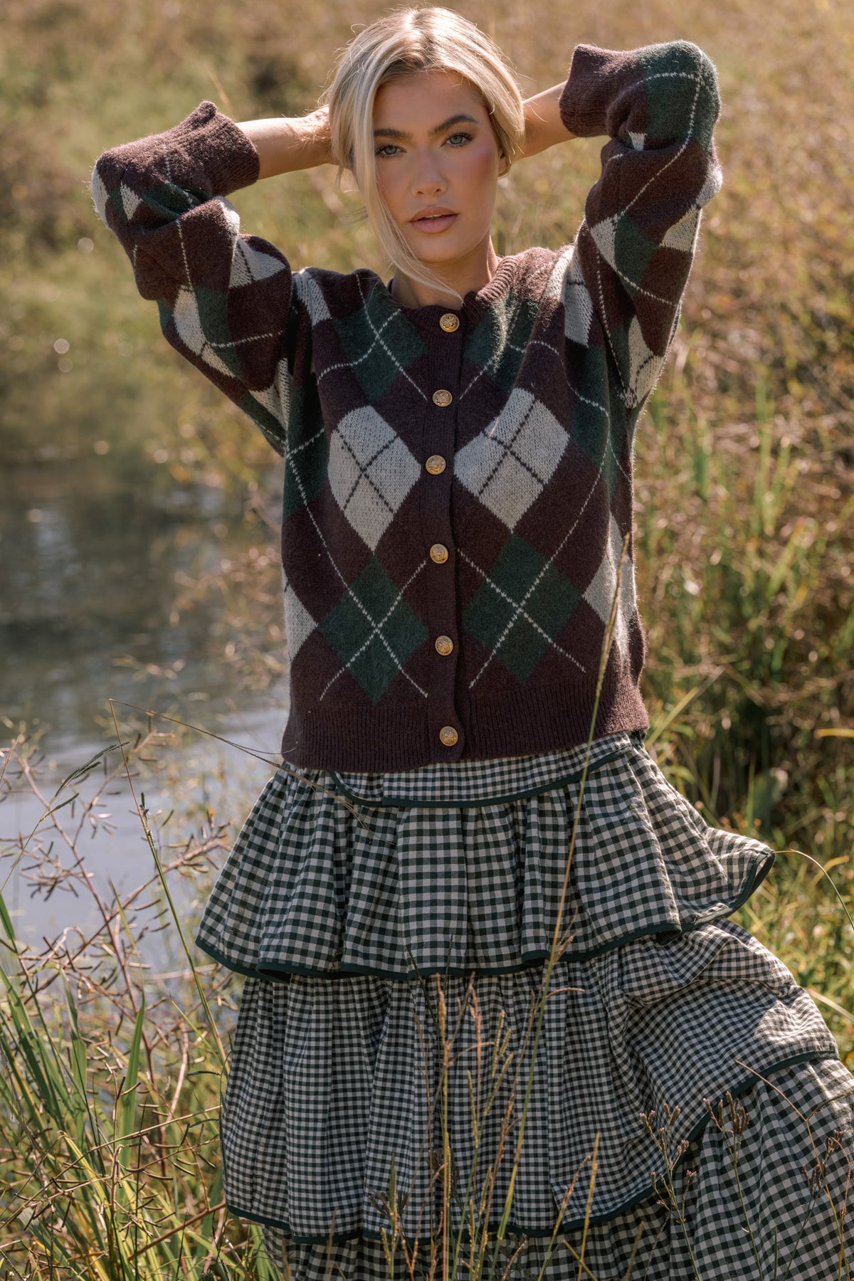 Woman wearing a brown argyle sweater with white, black, and green pattern details. She wears it over a green gingham dress while standing in front of a pond in a field. 