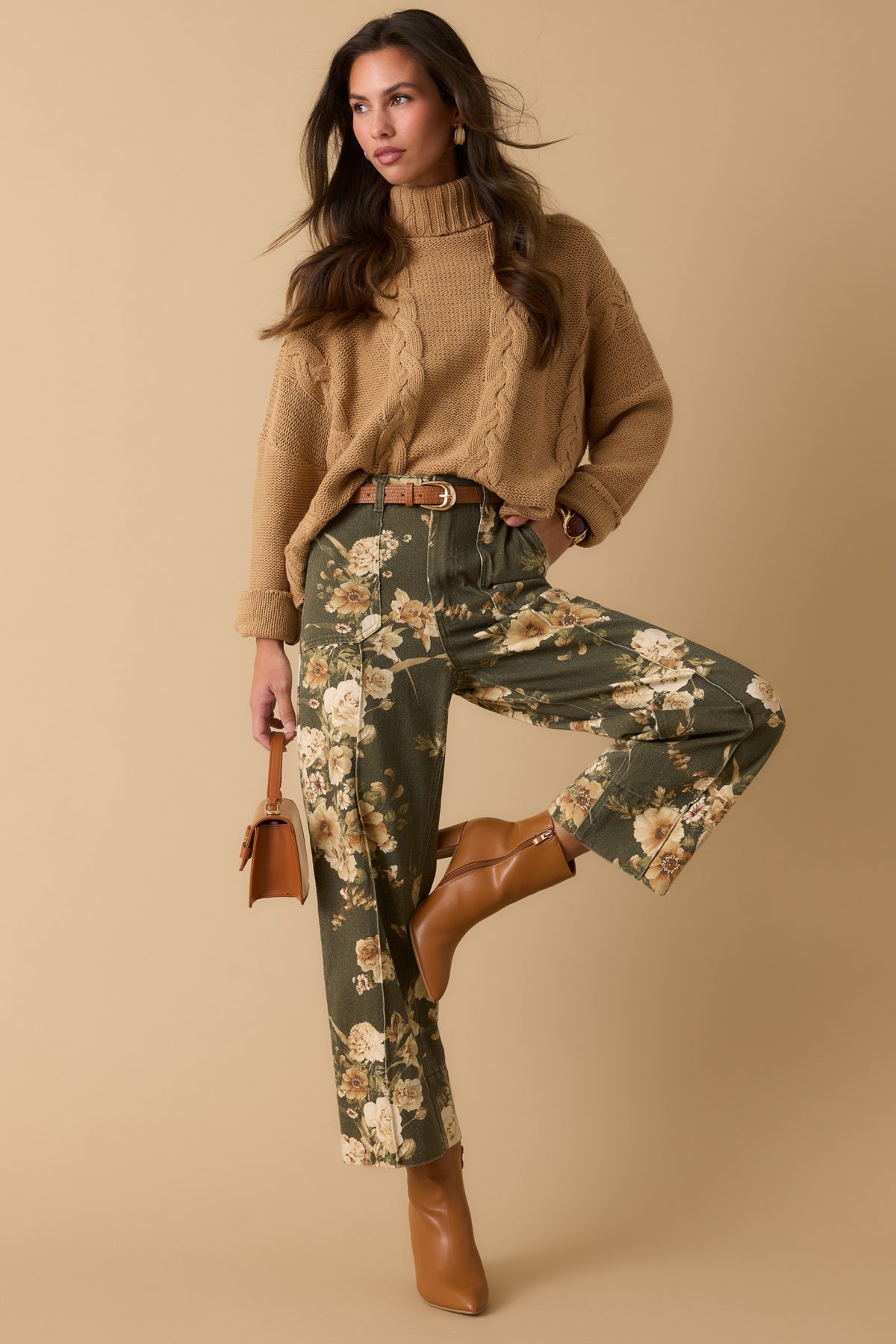 Woman in olive floral print pants, a camel cable knit turtleneck, with brown accessories against a beige backdrop. 