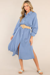 This blue dress features a collared neckline, functional buttons down the front, a front pocket on the left side of the bust, long sleeves with a cuff secured by a functional button, and two slits up the bottom hemline ending just below the knee.