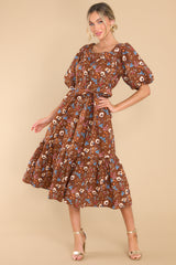 This brown floral dress features a square neckline, puffed sleeves, a self-tie belt, functional pockets, and a floral pattern.