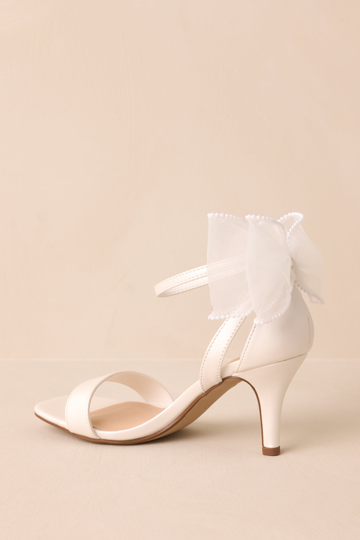 Three-quarter view of the white heels, showing the ankle strap with silver buckle and the detailed lace bow with pearl accents on the heel.