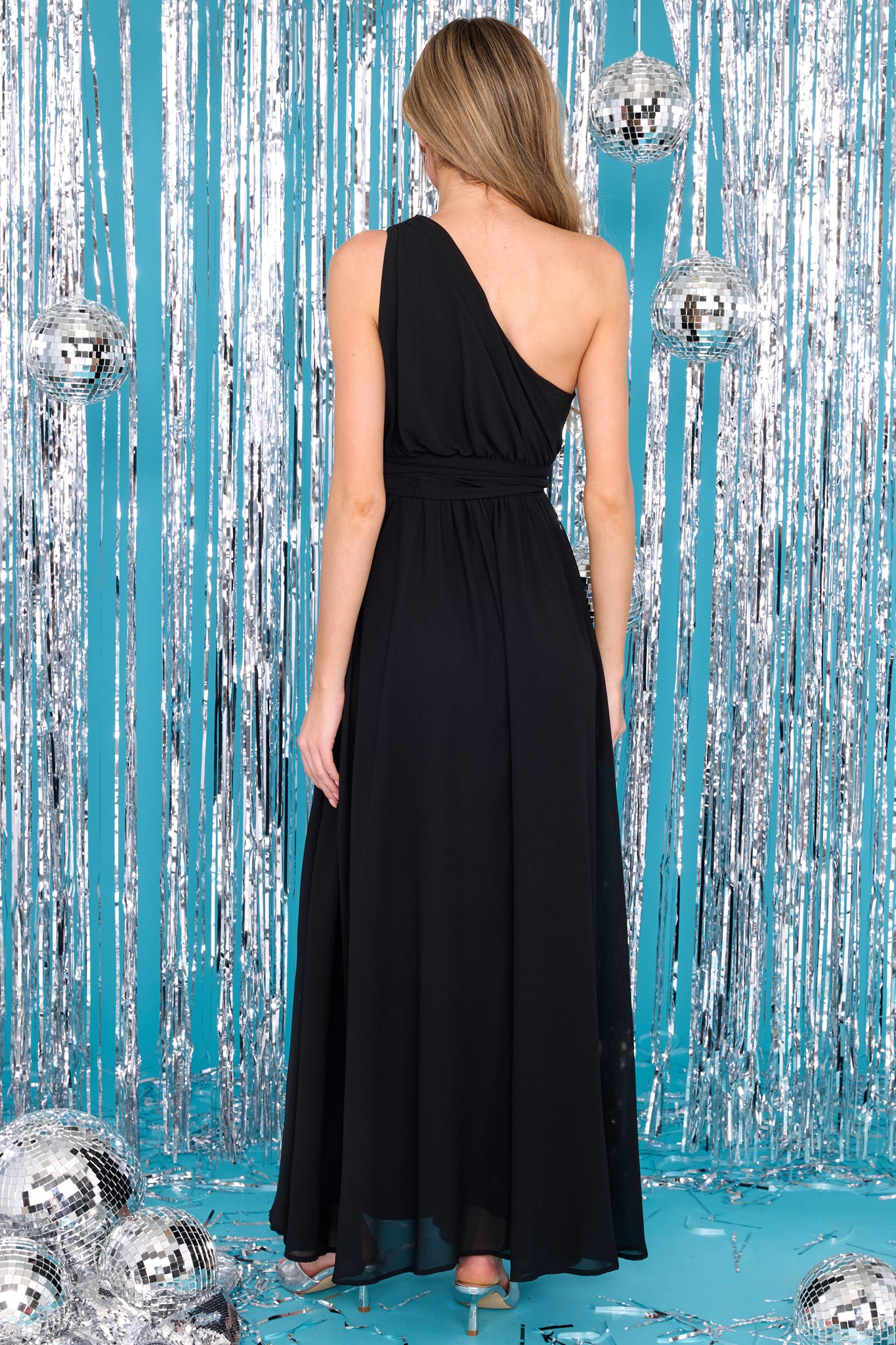 Back view of this dress that features a one-shoulder neckline with a non-slip rubber strip, a side zipper, an optional self-tie belt, a flowy skirt, and a slit up the leg.