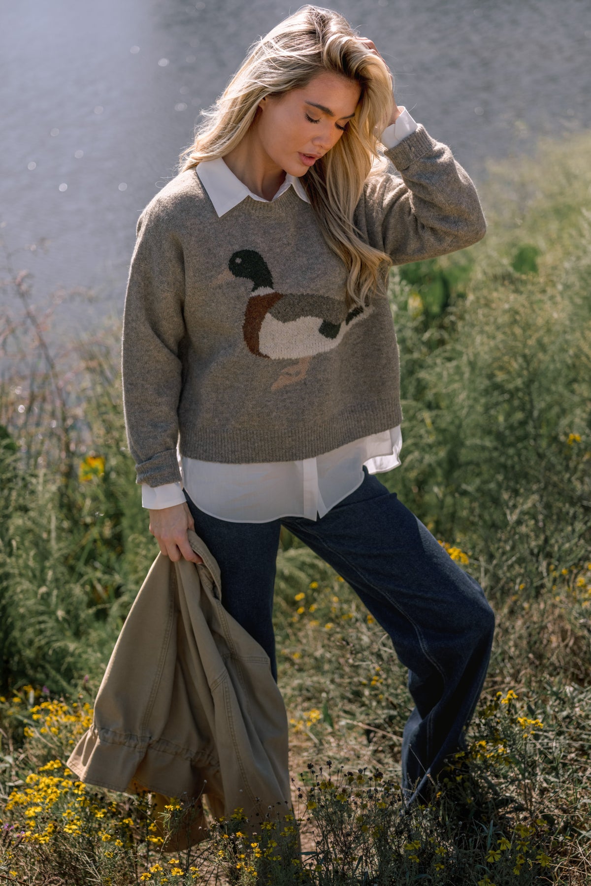 Woman modeling a taupe sweater featuring a duck on the front, a white button up shirt underneath, and dark jeans in front of a pond. She's holding a tan jacket in her hand, 