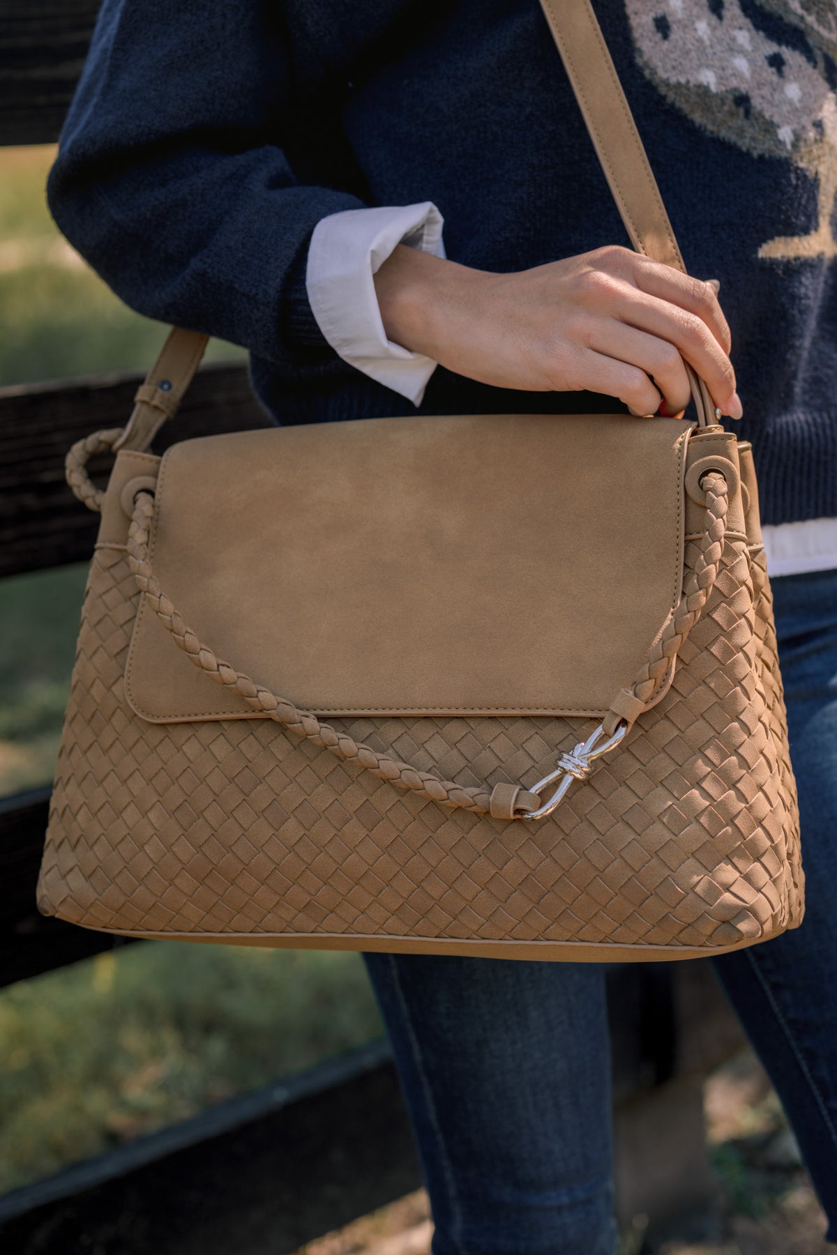 Tan satchel bag with rope detailing across the front being held by a model in front of a fence and field. 