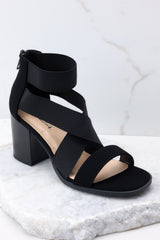 Outer-side view of these heels that feature a rounded toe, a strap across the top of the foot, two stretchy criss-cross straps, and a stretchy strap around the ankle, as well as a block heel and a zipper at the back of the ankle.