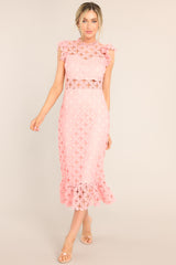 Fully view of this pink crochet lace midi dress featuring a high neckline with an illusion sweetheart neckline, ruffled shoulders, a peek-a-boo waistline which gives way into a sheath silhouette fit, and a flounced hem. 