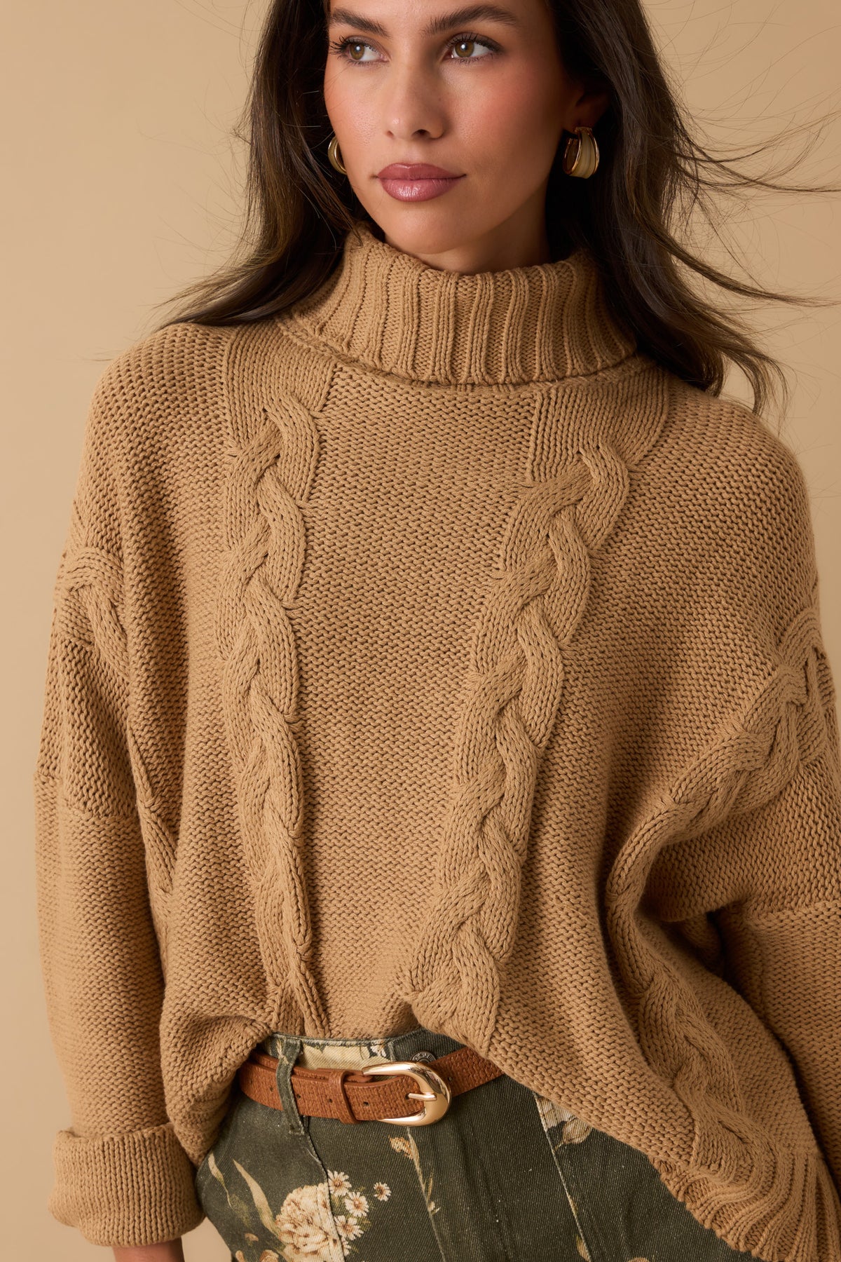 Woman wearing a camel colored, turtleneck sweater with a brown belt and olive floral print pants. 