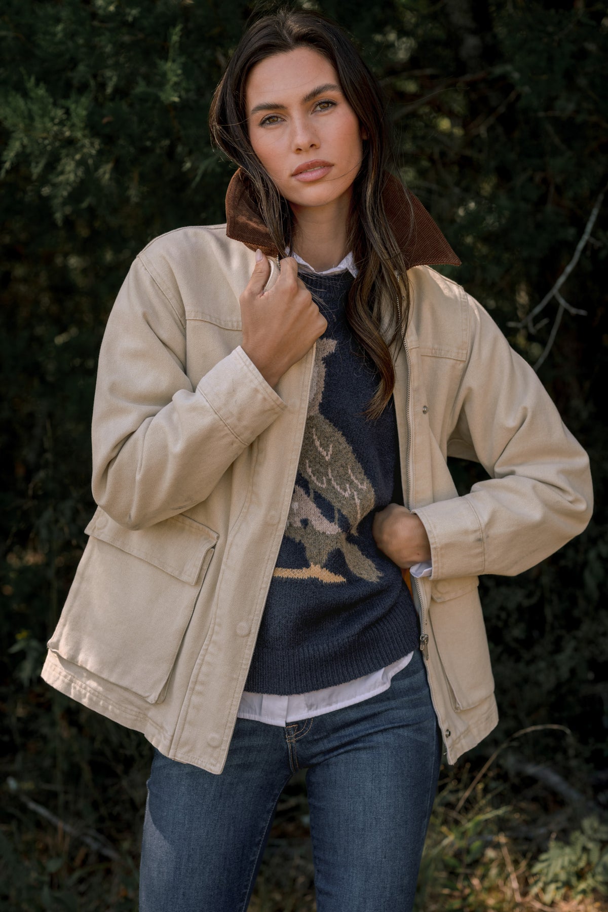 Woman modeling a light beige utility jacket with a brown corduroy collar, in front of dark green trees. 