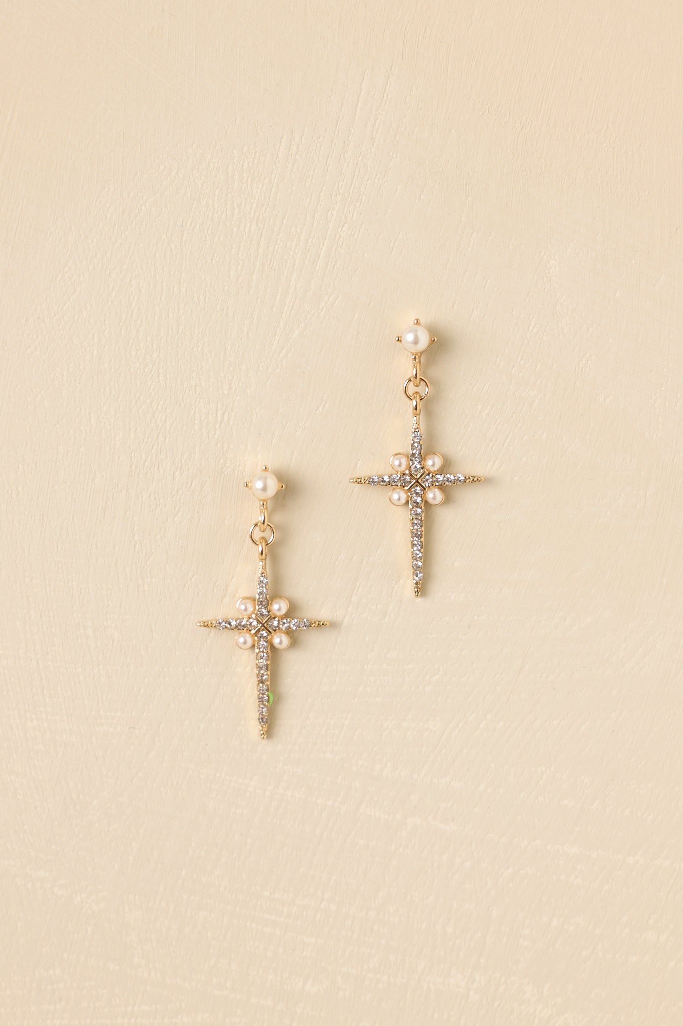 Gold drop earrings displayed on a beige background, showing the pearl studs, dangling crystal-accented cross, and post-back closure.