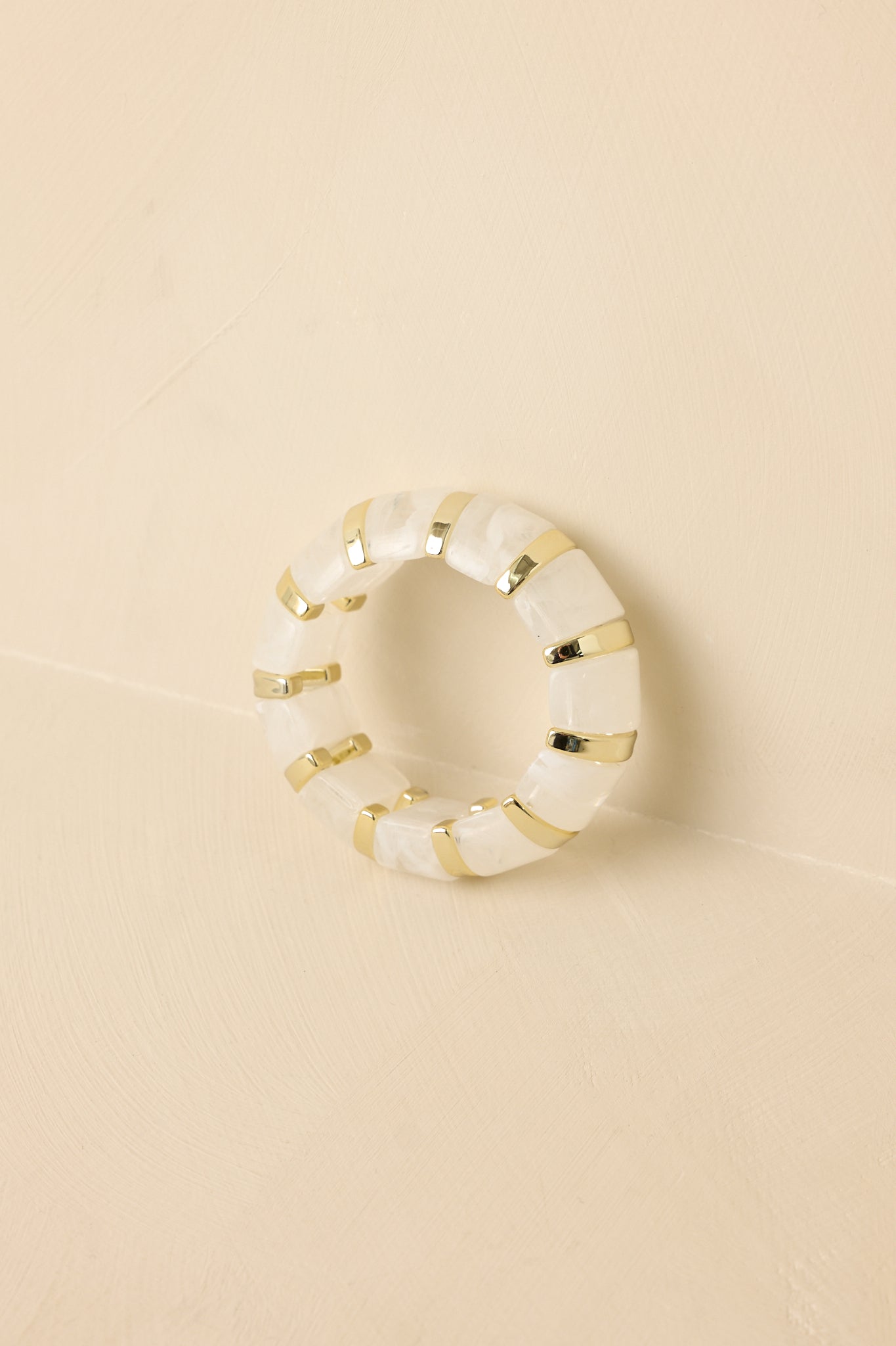 Gold-tone stretch bracelet featuring marbled white and clear beads set between polished rounded links against a beige backdrop.
