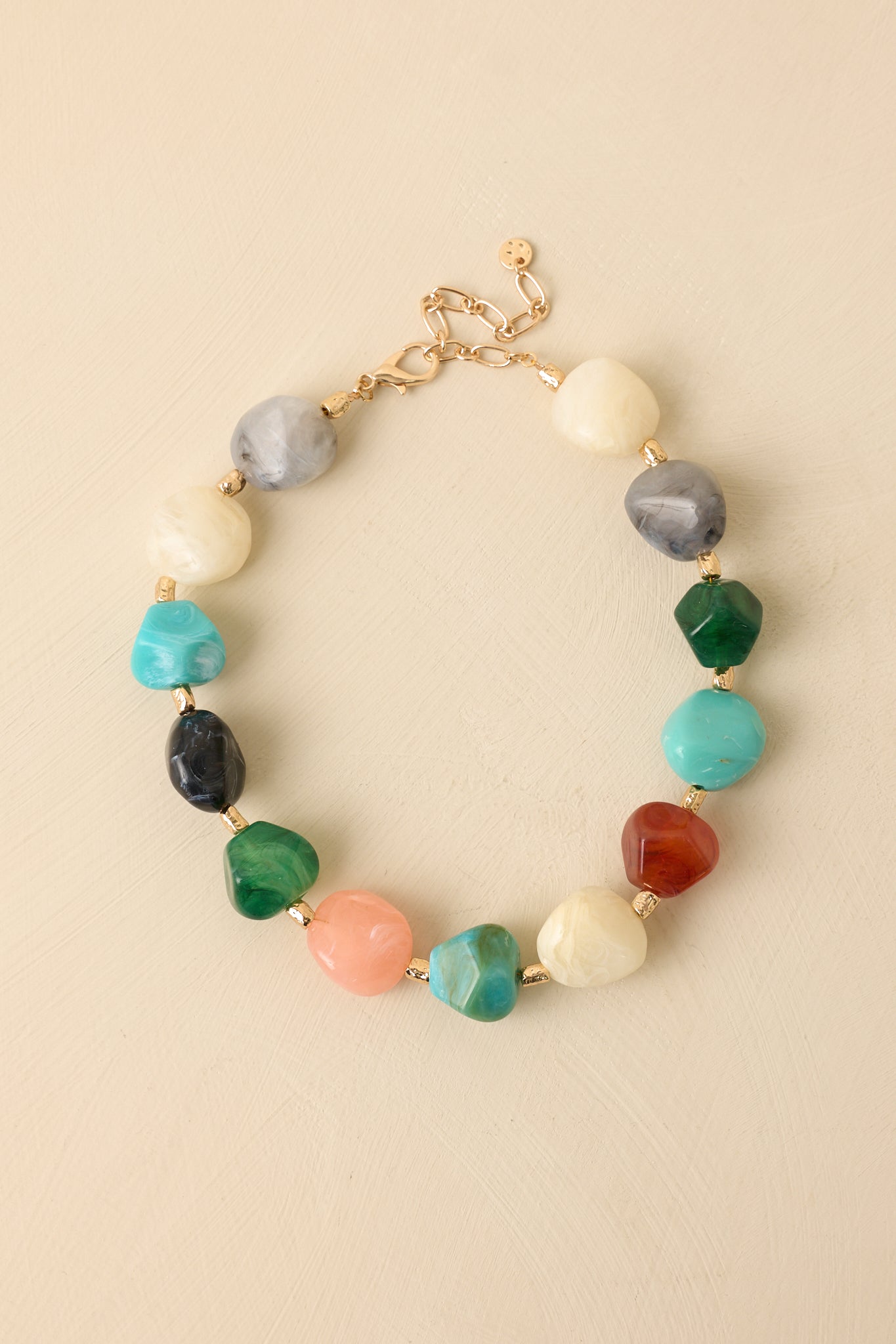 Multi-colored statement necklace displayed on a beige background showcasing its arranged gem-like stones and gold separators.