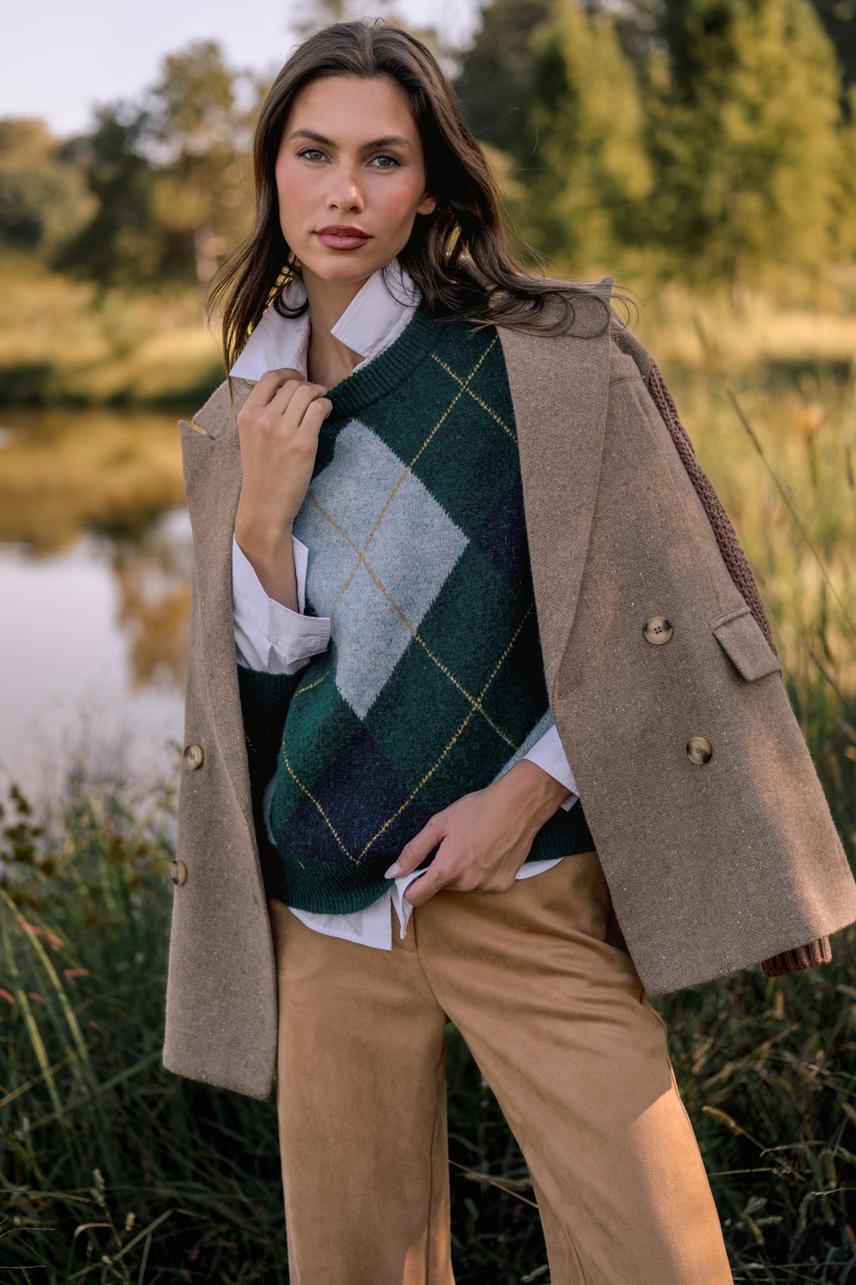 Woman wearing a tan blazer with knit sleeves over a green, ivory, and blue argyle sweater, with camel suede pants in front of a pond. 