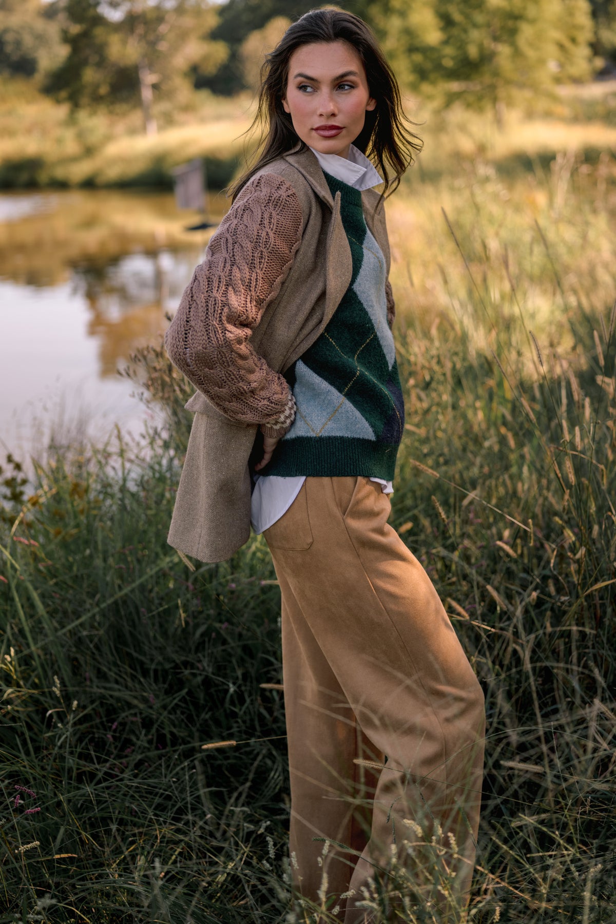 Brunette woman wearing a light brown blazer with knit sleeves over a green argyle sweater and camel suede pants while standing in front of a pond with tall grass. 