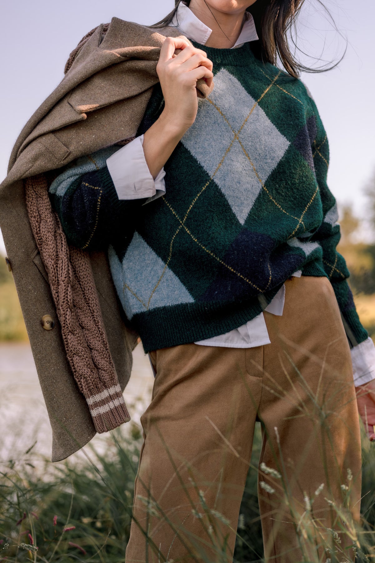 Woman wearing a green argyle sweater with light blue, navy, and gold details. It is paired with camel, suede pants and a light brown blazer slung over her shoulder while she stands in front of a pond. 