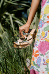 Angled view of these gold block heels that feature a square toe, two gold straps across the top of the foot, and a block heel.