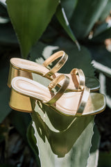 These gold block heels feature a square toe, two gold straps across the top of the foot, and a block heel.