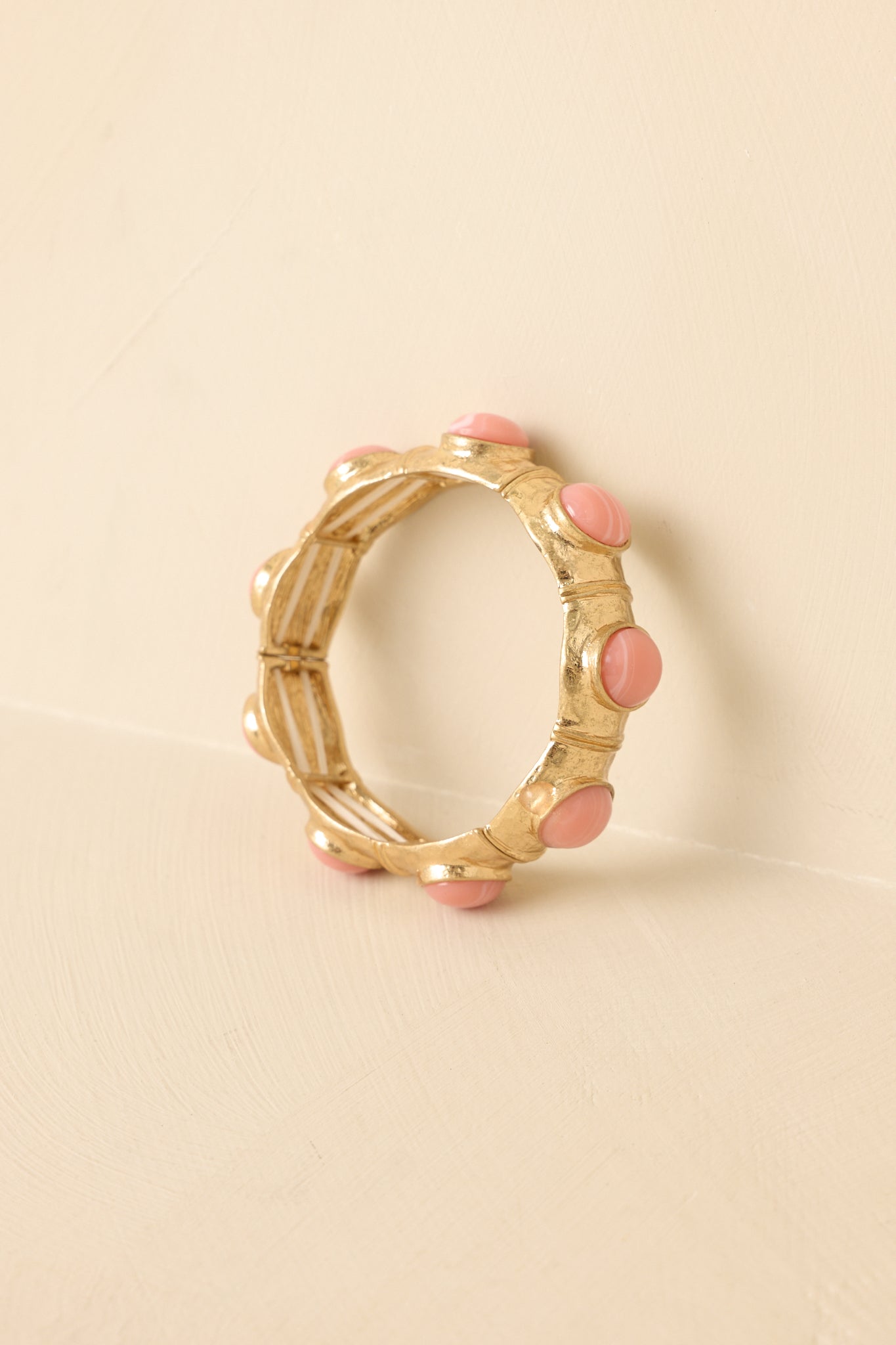 Sculpted gold-tone bracelet with soft pink cabochon accents on a beige backdrop, emphasizing its polished finish and bold shape.