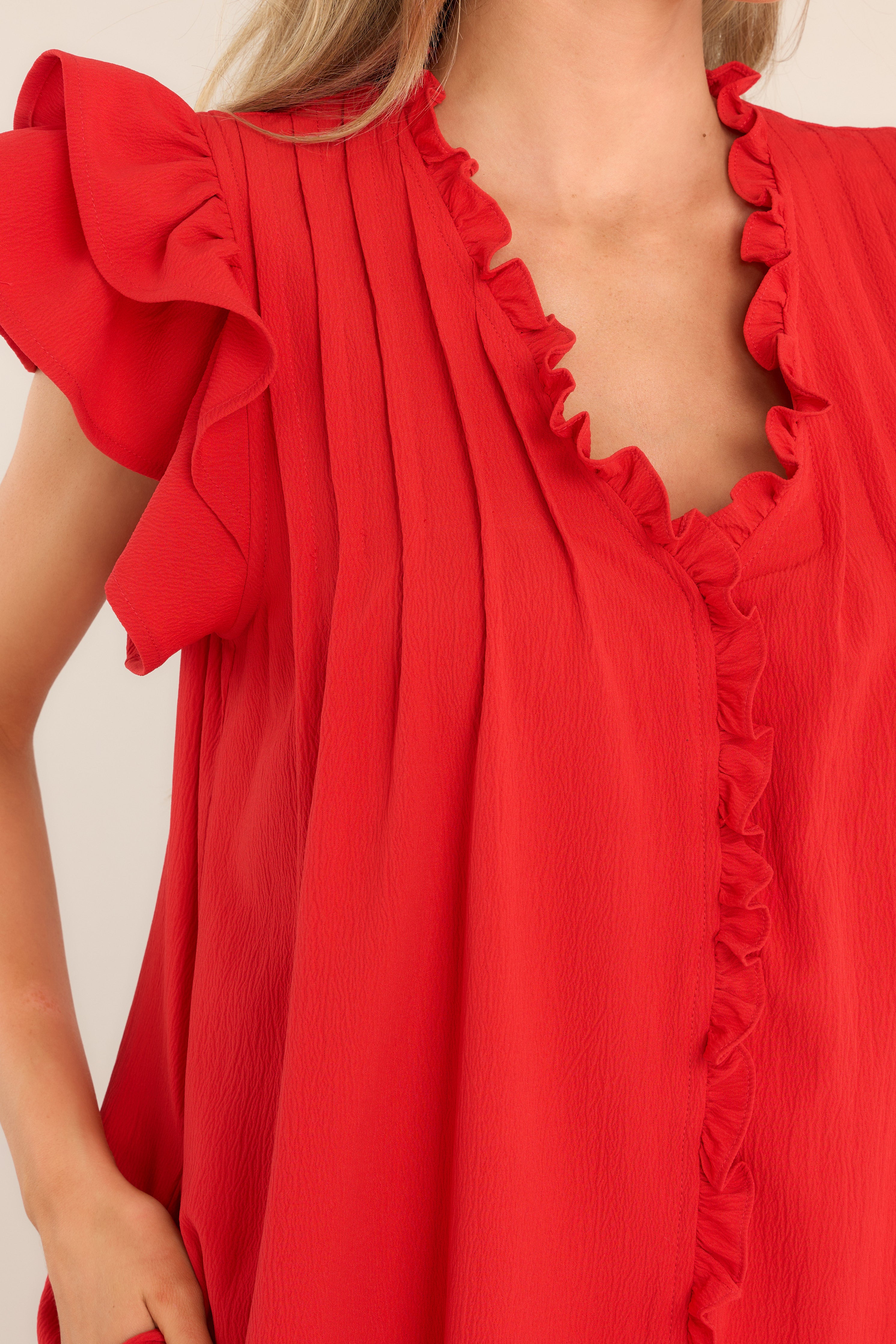 Close up view of this dress that features a ruffled v-neckline, a pleated chest, a full button front with ruffle detailing, functional hip pockets, and flutter sleeves. 