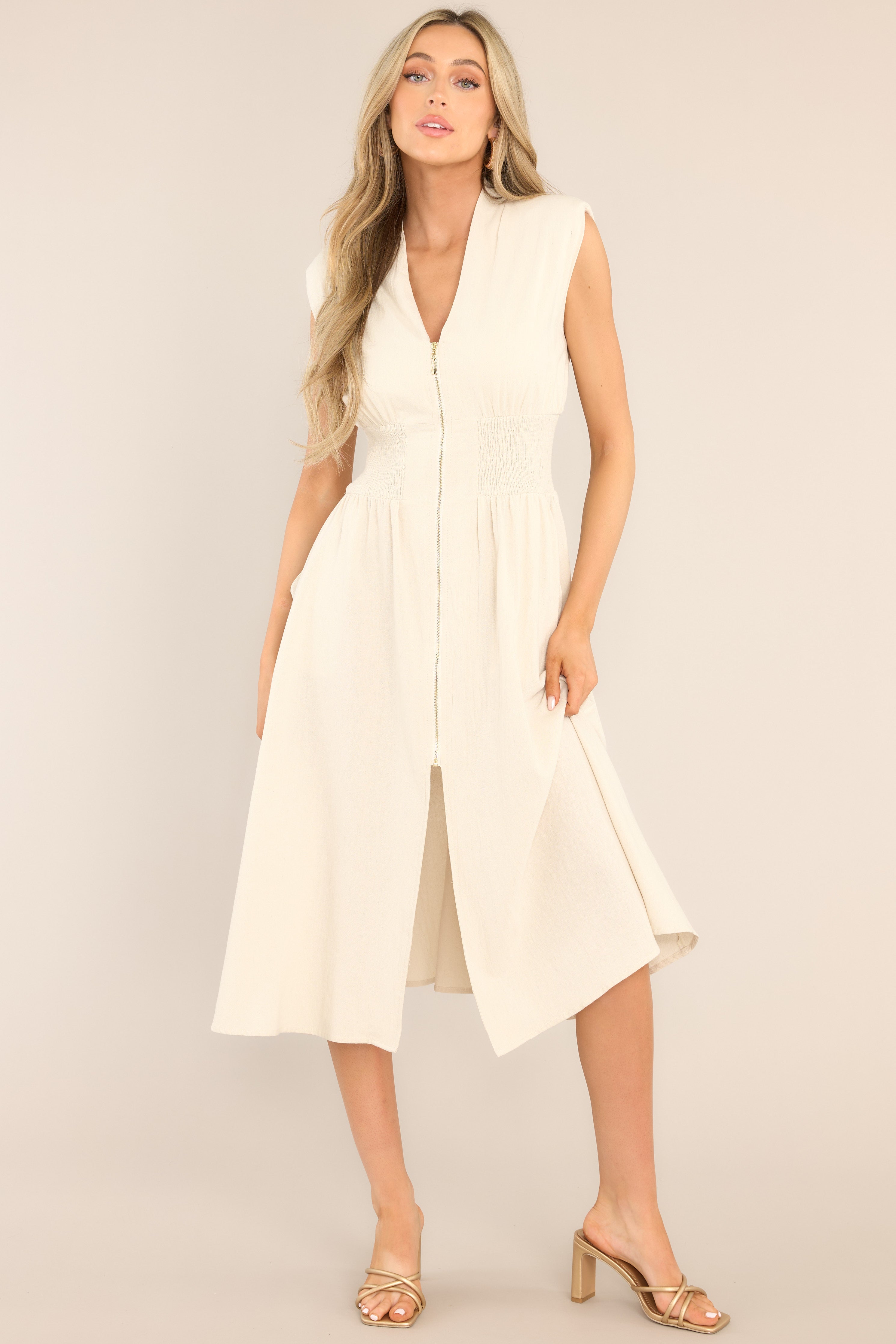 Full body view of this dress that features a deep v-neckline, shoulder padding, a functional zipper front, a fully smocked waist, functional pockets, and a front slit. 