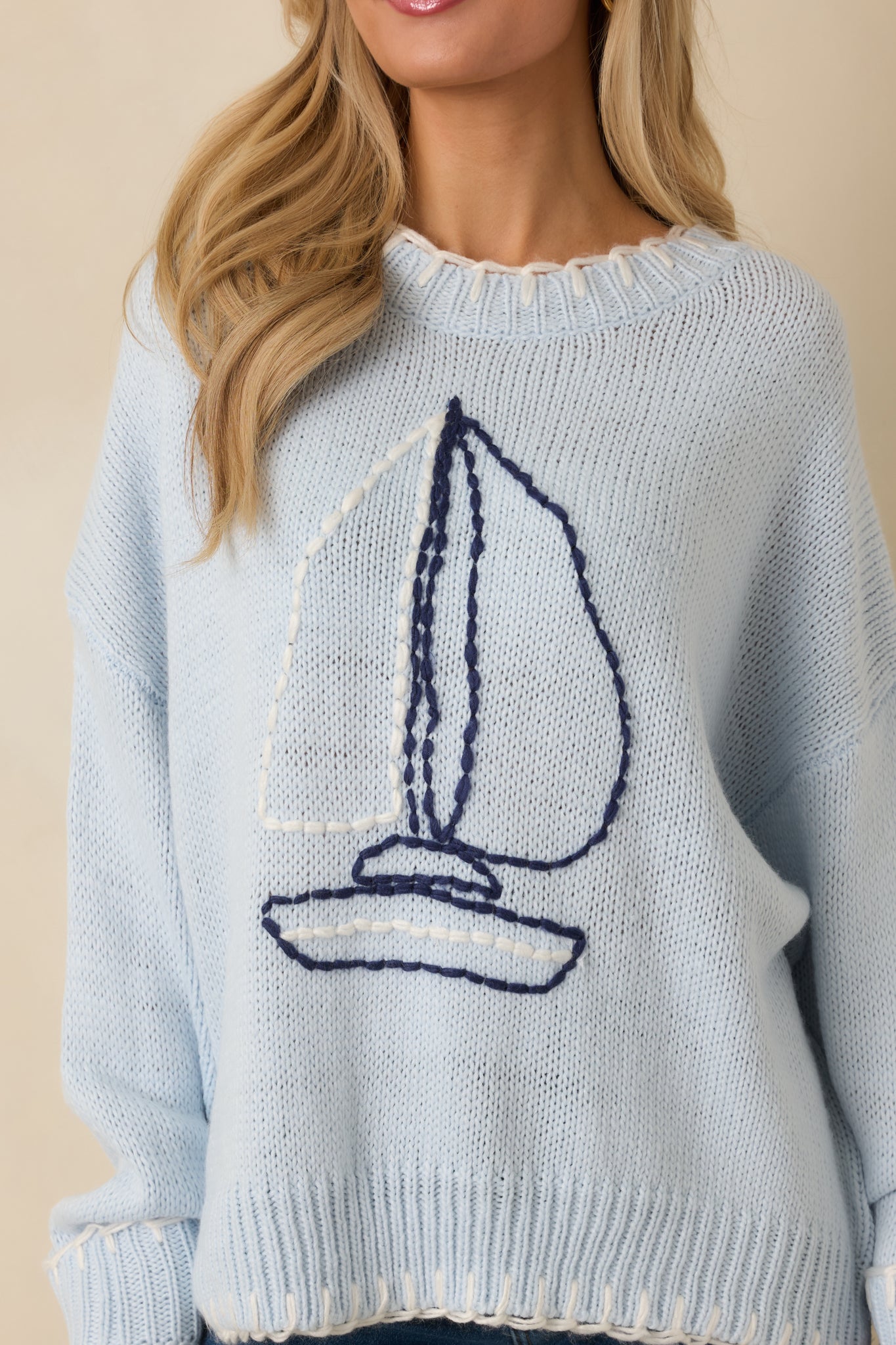 Close-up of the light blue knit sweater showing the embroidered sailboat detail and ribbed neckline.