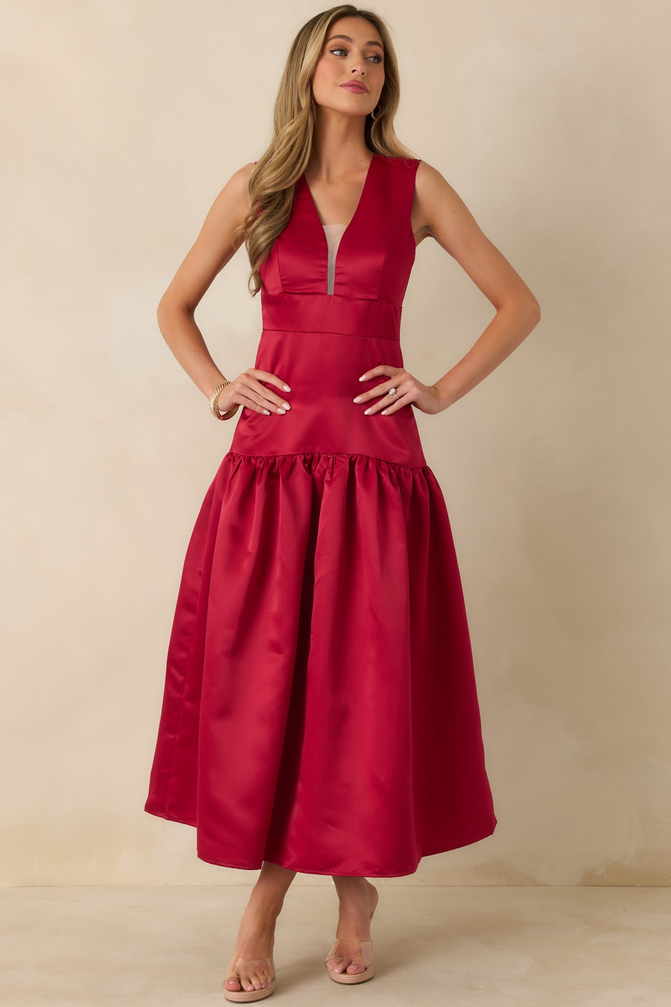 Red satin maxi dress with a fitted princess-seamed bodice, deep V-neckline with sheer insert, and a voluminous gathered skirt in a drop waist silhouette.