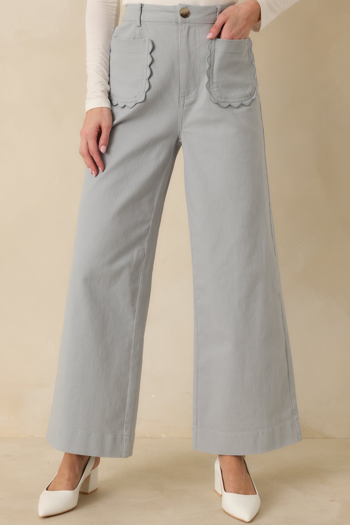 Dusty blue pants showing scalloped front patch pockets and smooth woven texture.
