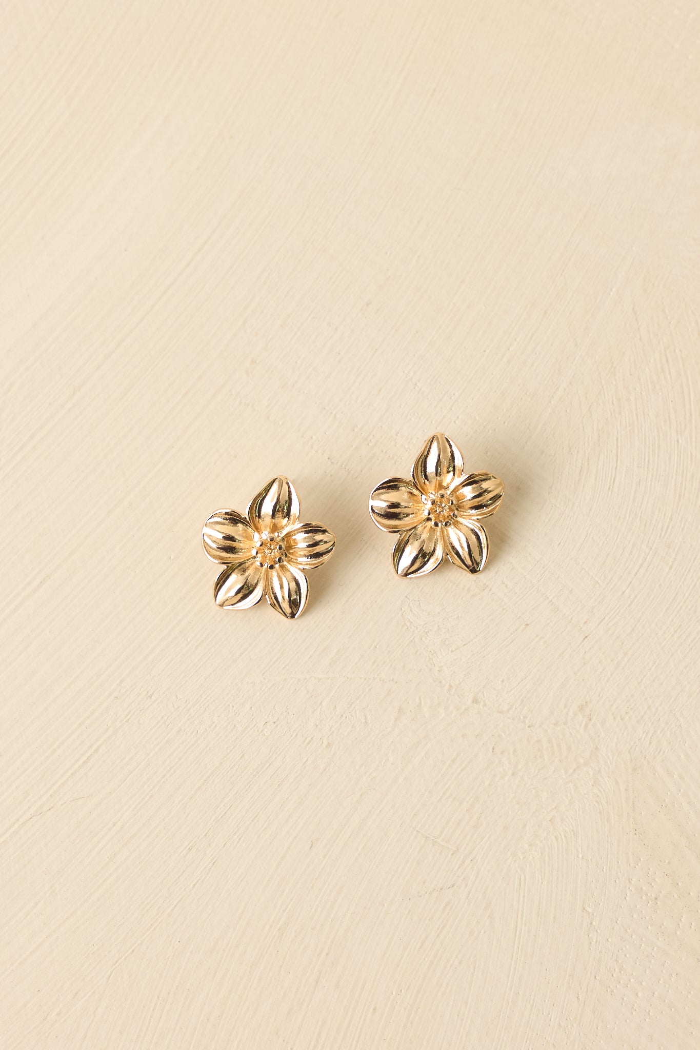 Front view of bold gold floral studs set against neutral beige, showing bloom design and refined craftsmanship.