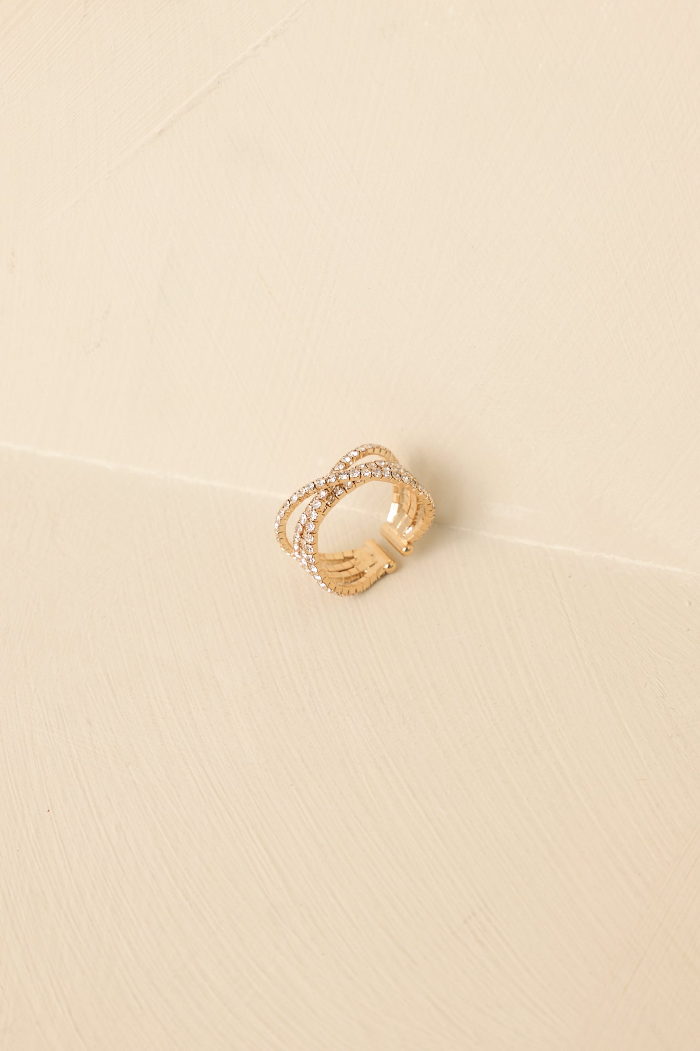 Open gold statement ring with intertwined bands and sparkling accents on a soft beige backdrop.
