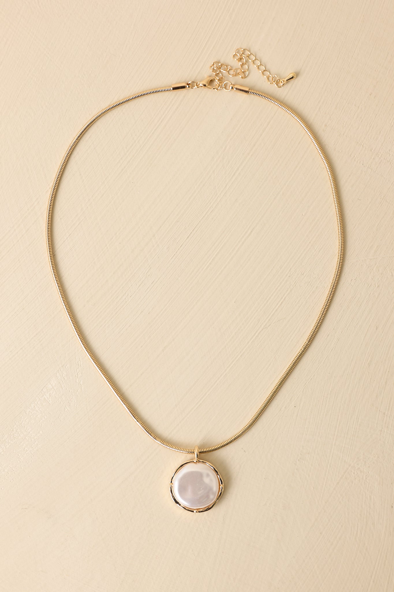 Gold and pearl pendant necklace displayed on a beige background, showing luminous pearl and polished gold frame.