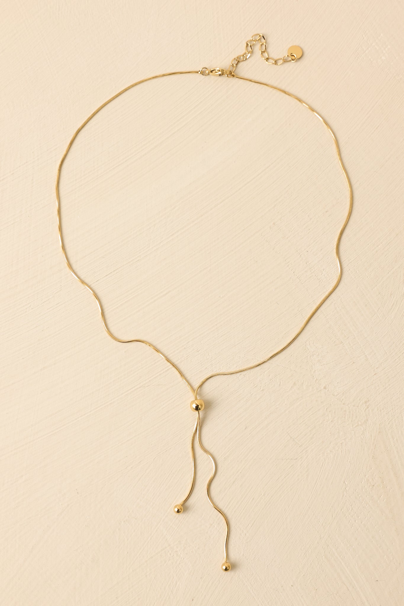 Gold lariat necklace displayed on a beige background, showcasing the fine chain and adjustable drop design.